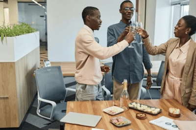 Three office colleagues smile as they clink champagne glasses together in celebration over a table with drinks and snacks.