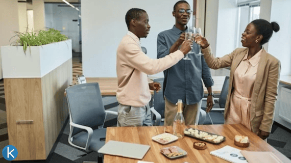 Three office colleagues smile as they clink champagne glasses together in celebration over a table with drinks and snacks.