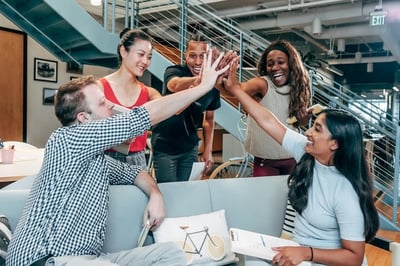 diverse group of five coworkers in a modern office high-fiving each other enthusiastically around a couch and coffee table.