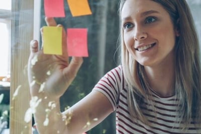 A woman peeling post-its with employee recognition quotes on them off a glass wall at work