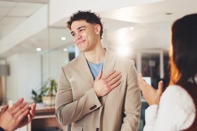 Man smiling with hand over heart as coworkers recognize him, showing how appreciation can reduce anxiety at work and build workplace confidence.