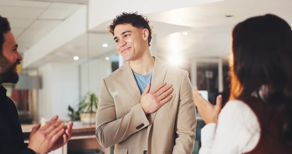 Man smiling with hand over heart as coworkers recognize him, showing how appreciation can reduce anxiety at work and build workplace confidence.