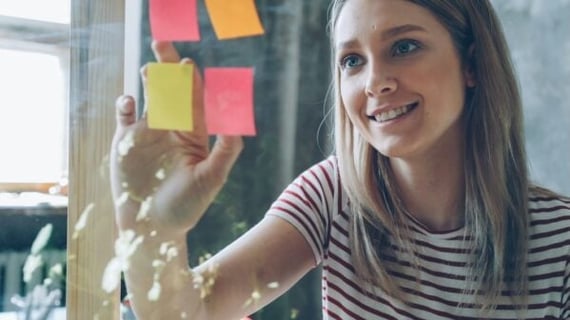 A woman peeling post-its with employee recognition quotes on them off a glass wall at work