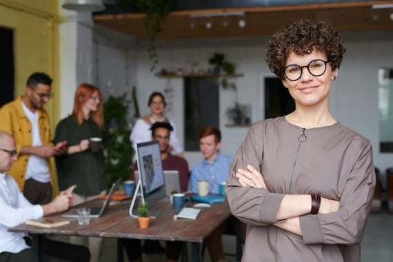 Smiling manager with her arms crossed in the foreground with employees working over a computer in the background