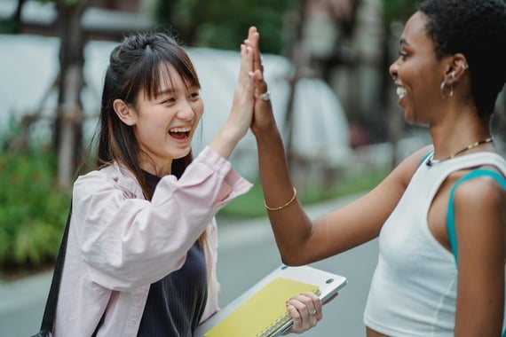 Two women high fiving outdoors while holding a laptop and notebooks