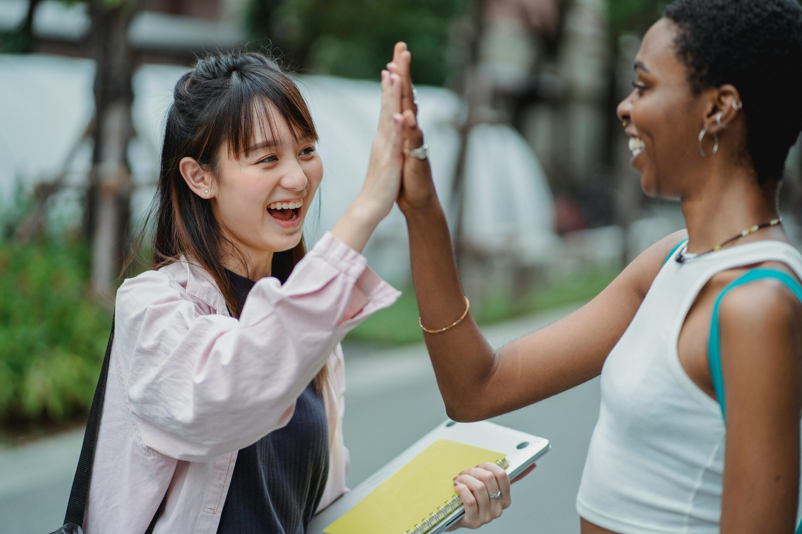 Two women high fiving outdoors while holding a laptop and notebooks