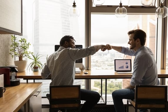 Two backlit men bump fists in front of work computers