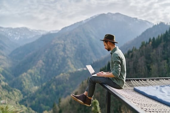 A man sitting on the edge of a cliff near the mountains with a laptop hopefully experiencing employee recognition