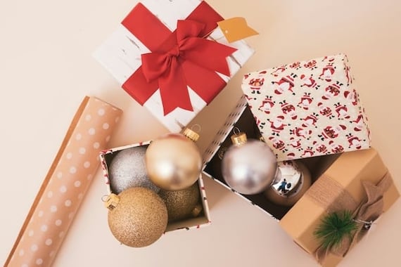 A festive tableau of holiday items, including giftwrap, a wrapped holiday gift, and ornaments, in gold, white, and red.