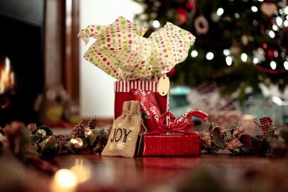 A festive holiday gift tableau, showing a cloth garland, wrapped gifts. A Christmas tree is shown twinkling faintly in the background.