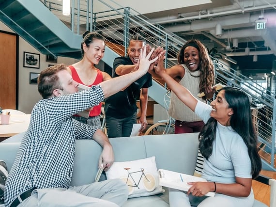 diverse group of five coworkers in a modern office high-fiving each other enthusiastically around a couch and coffee table.