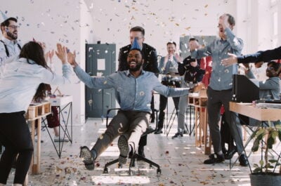 Employees wheeling person in office chair and giving birthday wishes to boss