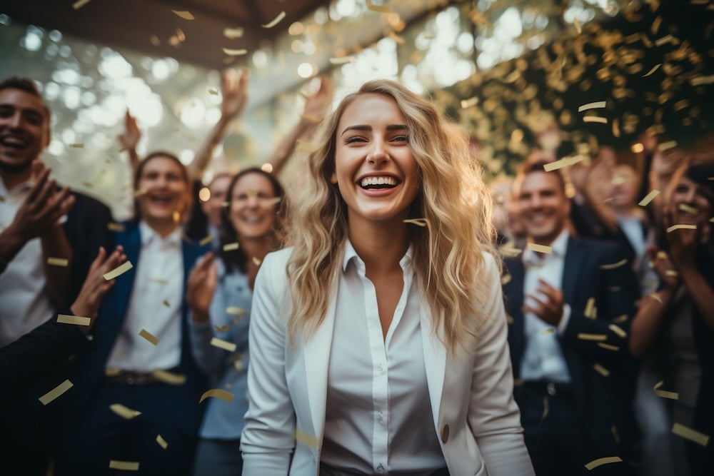 Employee recognition example with a happy employee in the foreground and blurred employees clapping in the background, plus gold confetti