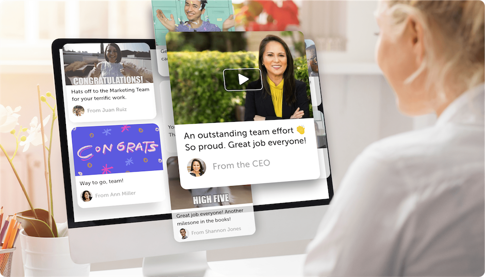 A person sits at a desk looking at a computer monitor displaying overlapping Kudoboard employee recognition post cards, including a “Congratulations!” note from Juan Ruiz, a “Congrats” graphic from Ann Miller, and a video message from the CEO applauding the team.