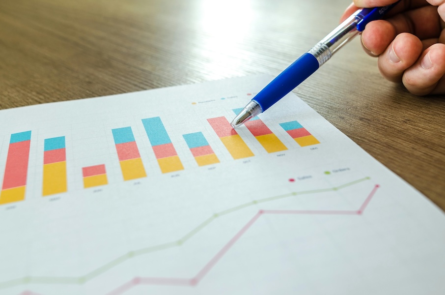 A hand holding a blue pen points at a printed chart of colorful stacked bar graphs and a line graph on a wooden desk surface, tracking employee recognition analytics.