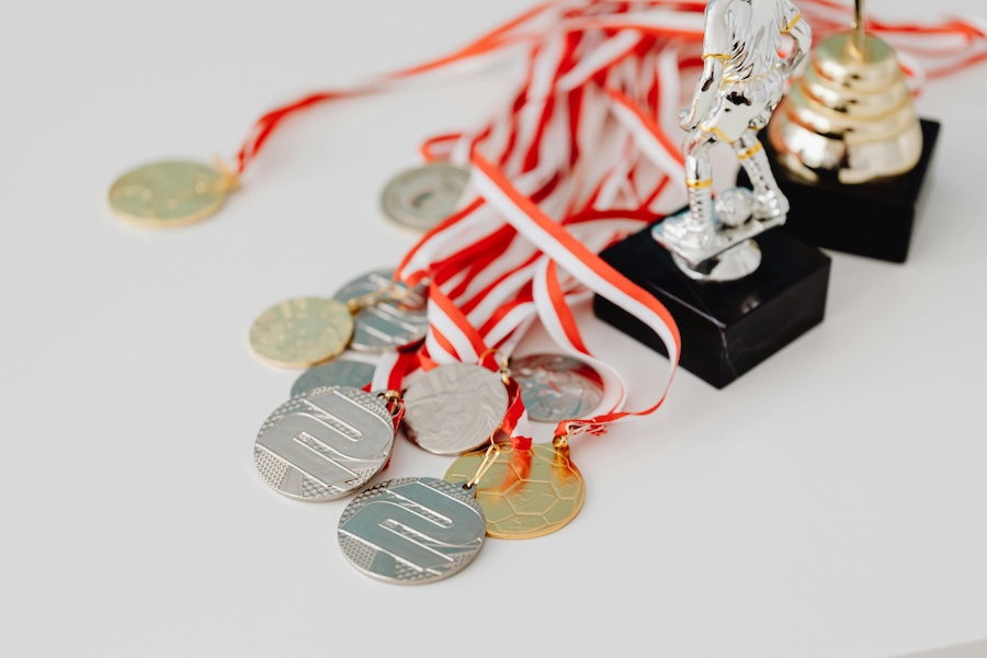 Assorted silver and gold medals on red-and-white striped ribbons alongside small sports trophies for employee recognition, all arranged on a white background.