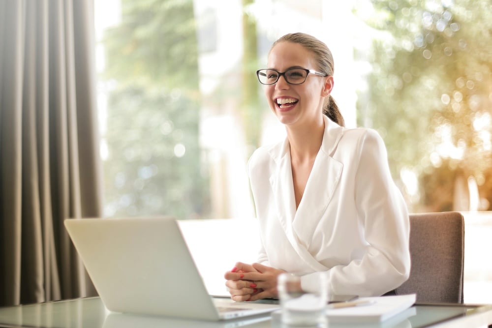 A professional woman with a ponytail in glasses and a white blouse is laughing. A computer, perhaps an ecard, is before her.