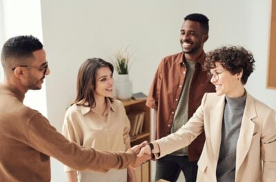 Four individuals in neutral clothing are smiling at one another. Two individuals in the foreground are shaking hands.