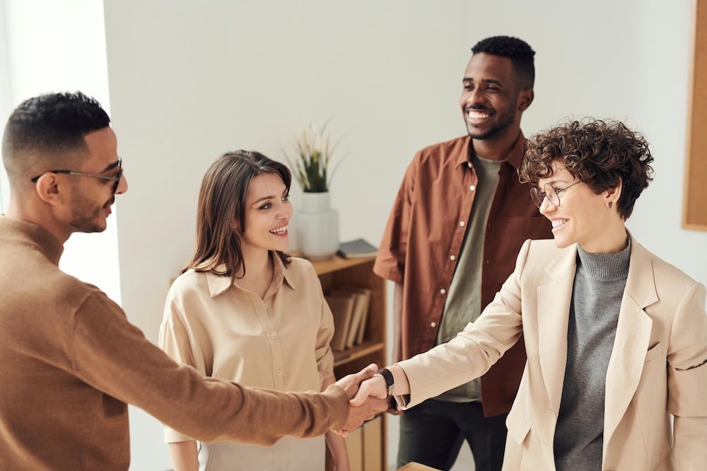 Four individuals in neutral clothing are smiling at one another. Two individuals in the foreground are shaking hands.