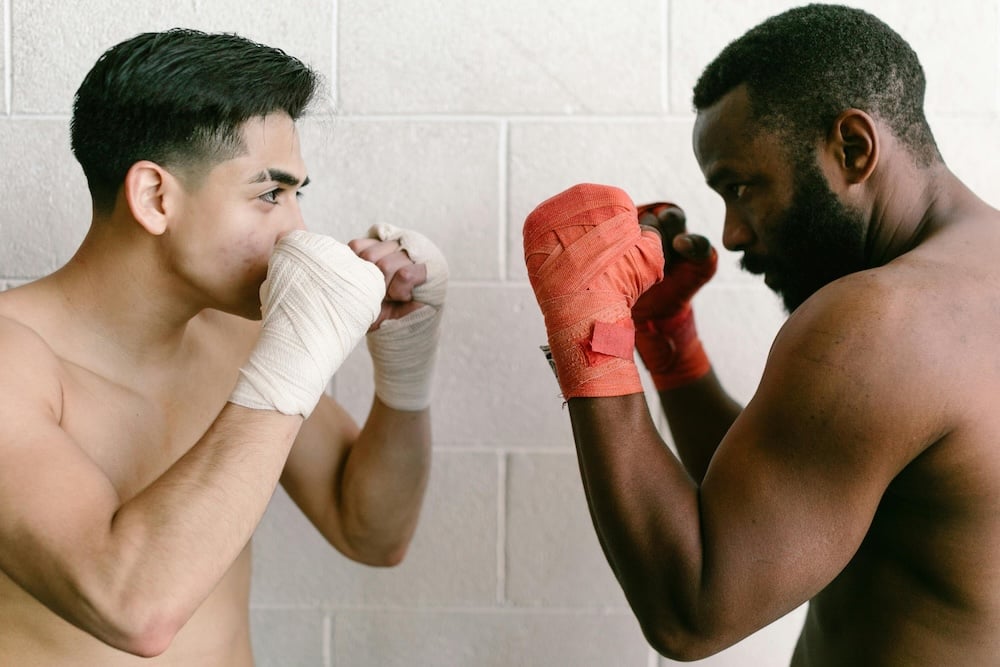 Two individuals square up in a boxing stance against one another. The one on the left has white glove-wraps, the one on the right has red.