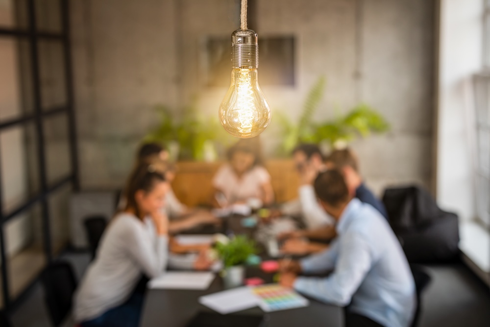 Six employees collaborating at a conference table in the background with a light bulb shining in the foreground