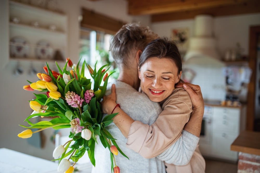 An adult daughter hugging her mom with a happy birthday wish and flowers
