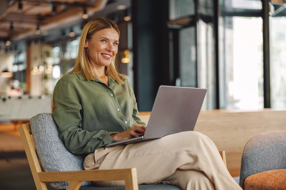 A woman in business casual clothes sitting and smiling at a laptop