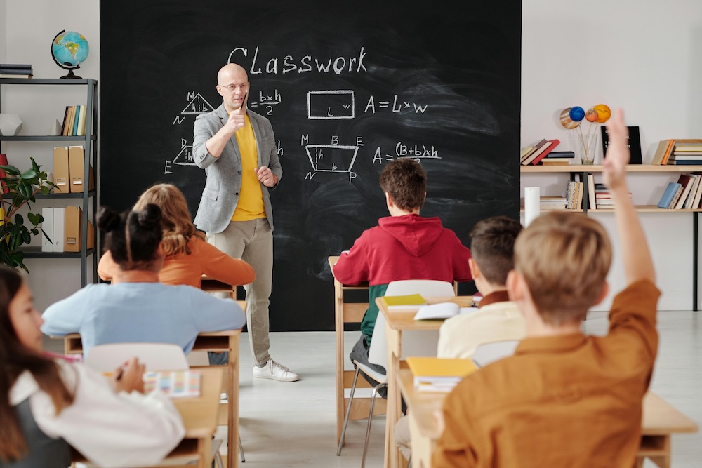 A teacher leads class in front of a chalkboard. In the foreground, a student raises their hand.