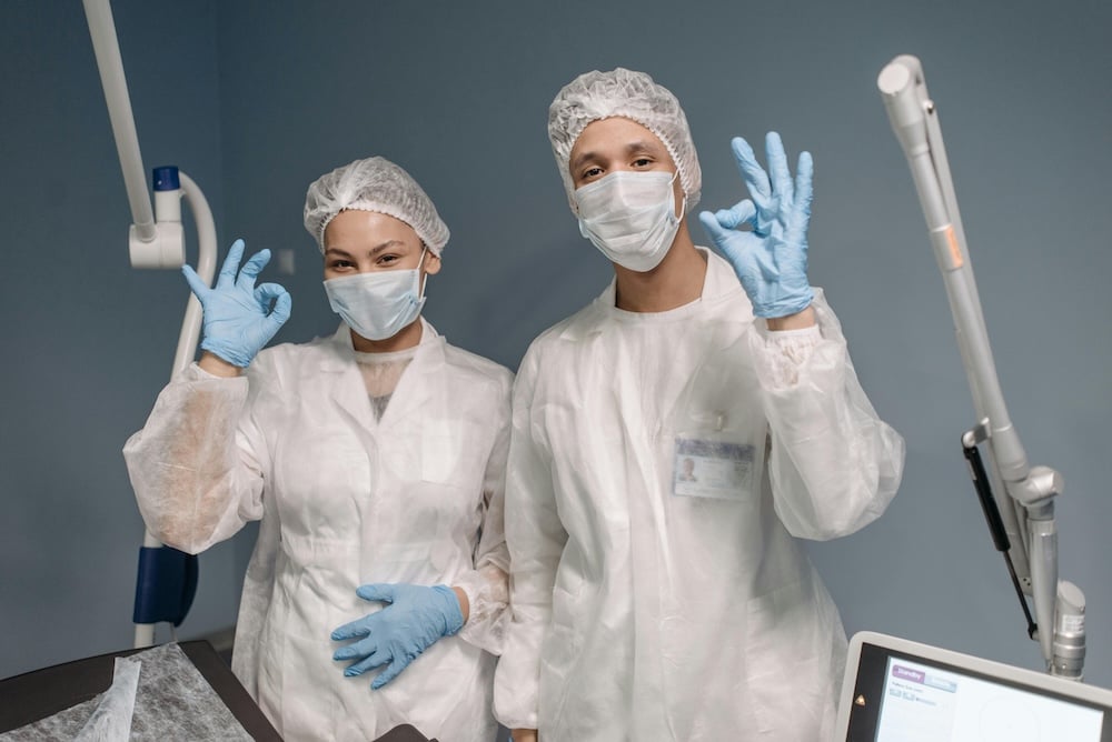 A pair of masked and suited-up surgeons pose at the camera, signaling 