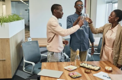 Three office colleagues smile as they clink champagne glasses together in celebration over a table with drinks and snacks.
