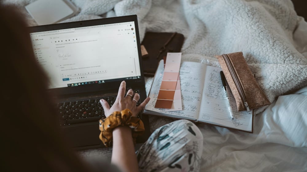 POV shot of a person sitting on their laptop on a bed with a journal open and paint samples