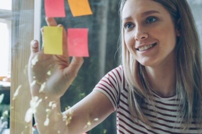 A woman peeling post-its with employee recognition quotes on them off a glass wall at work